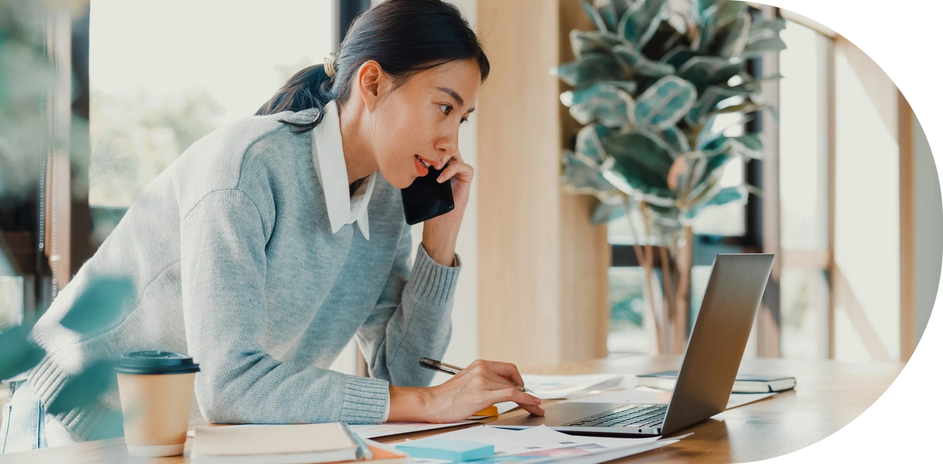 Mulher se concentra no computador e conversa ao telefone com colega